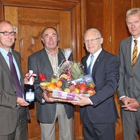 Im Amtszimmer stehen die vier Herren nebeneinander. Herr Sternath hält als Präsent eine Flasche Sekt in Händen. Werner Baader und OB Holzinger tragen gemeinsam einen großen Geschenkkorb.