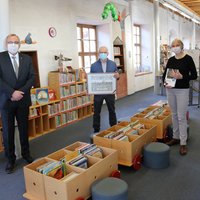 Die drei Personen sind in der Stadtbibliothek. Im Hintergrund sind Regalreihen mit Büchern zu erkennen. 