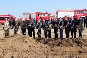 Das Bild zeigt die Personen in einer Reihe, jeder hat einen Helm auf dem Kopf und einen Spaten in der Hand. Im Hintergrund sind Feuerwehrfahrzeuge und mehrere Feuerwehrleute zu sehen.