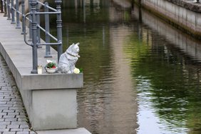 Die Skulptur aus der Ferne fotografiert mit Mäuerchen und Stadtbach
