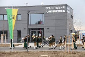 Eine Außenansicht des neuen Gebäudes. In großen Buchstaben steht "Feuerwehr Amendingen" an der Fassade, auf dem Weg vor dem Haupteingang zieht die Musikkapelle vorüber. Links im Bild weht die Amendinger Fahne vor dem Gebäude.