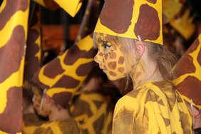 Auf dem Bild ist das Gesicht eines Mädchens der Blickfang, die als Giraffe geschminkt ist. Auf dem Kopf trägt sie einen spitzen Hut, der als Giraffenhals gestaltet ist. Um das Mädchen herum lauter Giraffen-Kinder.