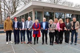 Die Gruppe ist dabei ein Band in Stadtfarben zu durchschneiden. Jeder hält dazu eine Schere in der Hand. Im Hintergrund ist das neue Gebäude von außen zu sehen, sie stehen vor dem Haupteingang.
