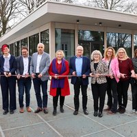 Die Gruppe ist dabei ein Band in Stadtfarben zu durchschneiden. Jeder hält dazu eine Schere in der Hand. Im Hintergrund ist das neue Gebäude von außen zu sehen, sie stehen vor dem Haupteingang.