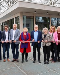 Die Gruppe ist dabei ein Band in Stadtfarben zu durchschneiden. Jeder hält dazu eine Schere in der Hand. Im Hintergrund ist das neue Gebäude von außen zu sehen, sie stehen vor dem Haupteingang.