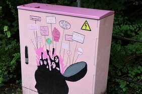 Ein rosaroter Stromkasten mit einem Kopf. Der Schädel ist aufgeklappt und darüber sind lauter Schilder, wie bei einer Demonstration. Die Aufschriften der Schilder wie im Fließtext beschrieben. 