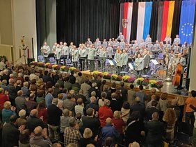 Das zahlreiche Publikum steht vor der Bühne, wo das Orchester ebenfalls steht und beklatscht wird. 