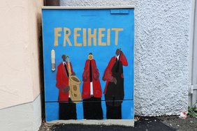 Auf einem blauen Stromkasten die Aufschrift "FREIHEIT", darunter drei Musiker mit Instrumenten.