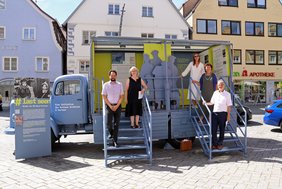 Ein hellblauer LKW mit abgerundeten Formen. Zwei Treppen führen auf die Ladefläche, die als Ausstellungsfläche dient. Auf den Stufen stehen die Personen.