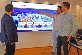 Die Personen stehen vor einem großen Monitor, auf dem das neue Startbild der Homepage zu sehen ist.