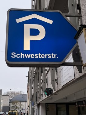 Im Bild ist das Leuchtschild vor dem Parkhauseingang abgebildet: ein großes P mit Dach auf blauem Grund, darunter der Schriftzug "Schwesterstraße". 