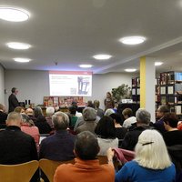 rund 30 sitzende Personen von hinten im Lesesaal des Stadtarchivs. Rechts Reihen von Bücherregalen. Vorne Stadtarchivar Engelhard, der eine Präsentation zeigt. 