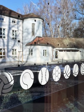 Unscharf im Hintergrund das gegenüber liegende Schulgebäude. Im Vordergrund scharf zu sehen ist ein Punktraster auf der Scheibe und stilisierte Bälle, mit denen im Gebäude viele Scheiben bedruckt sind.