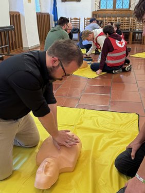 OB im Sitzungssaal auf einer Matte am Boden bei der Herzdruckmassage an einer Übungspuppe. Im Hintergrund eine andere Gruppe am Boden rund um eine Übungspuppe.