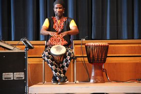 Der Trommler sitzt auf einem Stuhl und ist gerade dabei, mit den Händen auf die Trommel zu schlagen. Er hat einen sehr freundlichen Gesichtsausdruck.