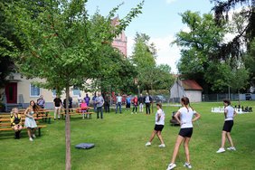 In einem großen Garten mit Apfelbäumen und vielen Büschen und Bäumen tanzen drei Mädchen. Die Gäste der Feier stehen und schauen zu.