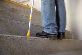 Zu sehen ist eine Treppe, die jemand hochsteigt. Von der Person sind nur die Schuhe zu sehen und ein Krückstock