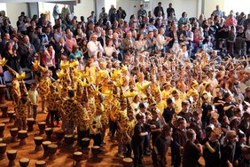 In der Mitte stehen viele Kinder in ihren Kostümen als Giraffen, Affen, Gazellen und Elefanten und tanzen. Rundherum die Zuschauer, die klatschen.