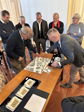 Zwei Männer sind am Tisch über ein Modell gebeugt, daneben drei weitere Modelle. Im Hintergrund stehen mehrere Personen, Mitglieder des Gestaltungsbeirats.