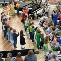Von der Empore aus fotografiert. Singende Kinder auf der Bühne, davor am Boden sitzen viele Kinder, auf den Stühlen aufmerksames Publikum.