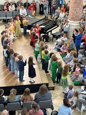 Von der Empore aus fotografiert. Singende Kinder auf der Bühne, davor am Boden sitzen viele Kinder, auf den Stühlen aufmerksames Publikum.