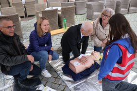 Die Gruppe am Marktplatz kniet auf dem Boden um eine Übungspuppe, die nur aus Oberkörper und Kopf besteht. AltOB Dr. Holzinger ist dabei, das Herz der Puppe zu drücken, die anderen schauen zu und unterhalten sich.
