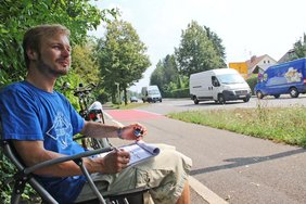 Urs Keil sitzt auf einem Campingstuhl am Gehsteig. Ein Grünstreifen trennt ihn vom viel befahrenen Bayernring. Er hat ein Handzählgerät in der Hand und ein Klemmbrett mit Tabellen auf dem Schoß