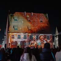 Am Haus der Kramerzunft wird ein Video abgespielt, im Bild sind mittelalterliche Bauern versammelt, dazu eine historische Fassade des Gebäudes.