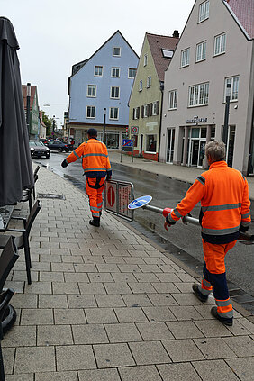 Zwei Bauhof-Mitarbeiter tragen ein Schild weg. 