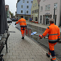 Zwei Bauhof-Mitarbeiter tragen ein Schild weg. 