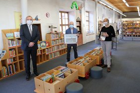 Die drei Personen sind in der Stadtbibliothek. Im Hintergrund sind Regalreihen mit Büchern zu erkennen. 