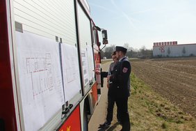 Zwei Feuerwehrmänner stehen vor den Bauplänen, die an einem Fahrzeug befestigt sind. Im Hintergrund gibt der Baumarkt Bauhaus Orientierung, wo das Fahrzeug steht.