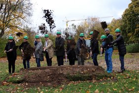 Klassisches Spatenstich-Bild: Eine Erdhaufen und dahinter die Personen mit Helm und Spaten.