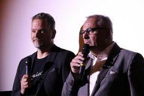 Die beiden Herren stehen nebeneinander im Spotlight vor der Kinoleinwand.