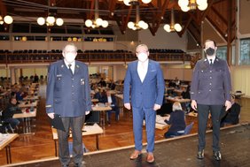 Die drei Männer stehen nebeneinander auf der Bühne der Stadthalle. Im Hintergrund ist der Saal zu sehen, einige Stadträtinnen und Stadträte jeweils einzeln an einem Tisch. Der Hintergrund ist etwas in Unschärfe gesetzt, um die Aufmerksamkeit auf die drei Hauptpersonen zu konzentrieren.