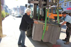 Zu sehen ist das am Tieflader mittels Bändern befestigte Modell, das wenig über dem Boden schauckelt und von den beiden Männern auf den richtigen Standort geschoben, bzw. gezogen wird.