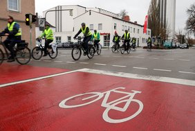 Eine Gruppe fahrender Radfahrer mit gelben Warnwesten wird gezeigt, im Vordergrund eine Markierung auf der Straße, die ein Fahrrad abbildet.