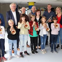 Die Kinder stehen in der ersten Reihe mit an den Mund angelegten Trompeten, die weiteren Personen in der Reihe dahinter. 