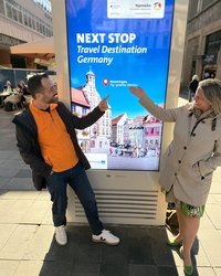 Die beiden Personen deuten auf ein digitales Plakat mit der Aufschrift: "Next Stop Travel Destination Germany: Memmingen"; Grafisch zeigt das Plakat den Marktplatz mit Brunnen und Rathaus.