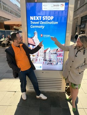 Die beiden Personen deuten auf ein digitales Plakat mit der Aufschrift: "Next Stop Travel Destination Germany: Memmingen"; Grafisch zeigt das Plakat den Marktplatz mit Brunnen und Rathaus.