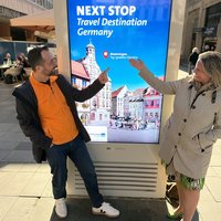 Die beiden Personen deuten auf ein digitales Plakat mit der Aufschrift: "Next Stop Travel Destination Germany: Memmingen"; Grafisch zeigt das Plakat den Marktplatz mit Brunnen und Rathaus.