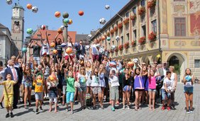 Die Jugendlichen werfen ihre Bälle in die Luft. Die Gruppe mit Schülern und Lehrern steht vor dem Brunnen am Marktplatz