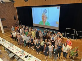 Perspektive von oben: die Personengruppe, darüber auf einer Leinwand die Ministerin. 