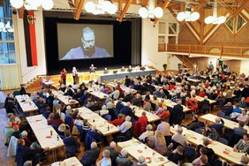 Wieder Blick von oben (von der Empore) auf den Saal: die Gäste sitzen an langen Tischreihen, viele haben Getränke vor sich und die Willkommentaschen liegen vor ihnen auf dem Tisch. Vorne links die Gebärdendolmetscherin, die zu den Gehörlosen vor sich übersetzt. Der OB ist auf der Leinwand zu sehen, er selbst steht an die Bühne gelehnt auf dem Vorpodest mit dem Podium. Auf dem Podium die Referatsleiter. 