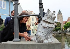 Die weiß lasierte Katzenfigur thront auf einem Mauerabsatz, die Künstlerin Cornelia Brader ist hinter dem Kunstwerk durch das Geländer zum Stadtbach zu erkennen. Im Hintergrund der Stadtbach und der Turm der Frauenkirche