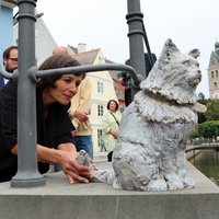 Die weiß lasierte Katzenfigur thront auf einem Mauerabsatz, die Künstlerin Cornelia Brader ist hinter dem Kunstwerk durch das Geländer zum Stadtbach zu erkennen. Im Hintergrund der Stadtbach und der Turm der Frauenkirche