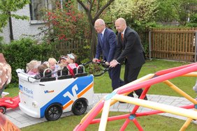 Im Vordergrund ist ein Klettergerüst zu sehen. Dahinter schieben OB Holzinger und Herr Schinke schwungvoll den großen Kinderwagen um die Kurve.