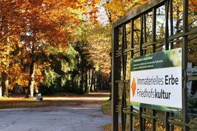 Das Schild ist groß an der geöffneten Tür im Vordergrund, unscharf dahinter ein Blick auf goldenes Herbstlaub der Friedhofsbäume
