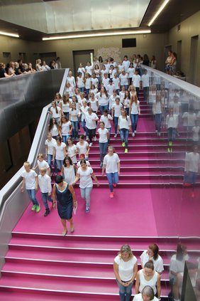 Zu sehen sind weiß gekleidete Chorsängerinnen und Sänger, die das pinkfarbende Treppenhaus hinabsteigen.