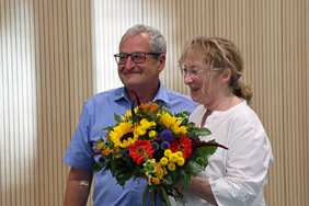 Sie hält einen Blumenstrauß in Händen, beide schauen nach links in die gleiche Richtung und lächeln.
